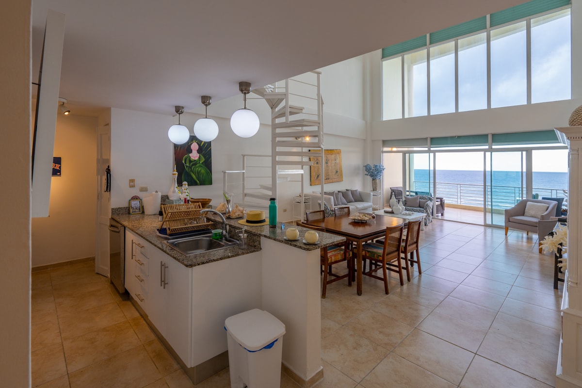 An open-concept living space features a modern kitchen with a granite countertop and pendant lighting. A dining area with wooden chairs is positioned beside floor-to-ceiling windows, offering views of the ocean. Light-colored furnishings create a bright and airy atmosphere throughout the room.