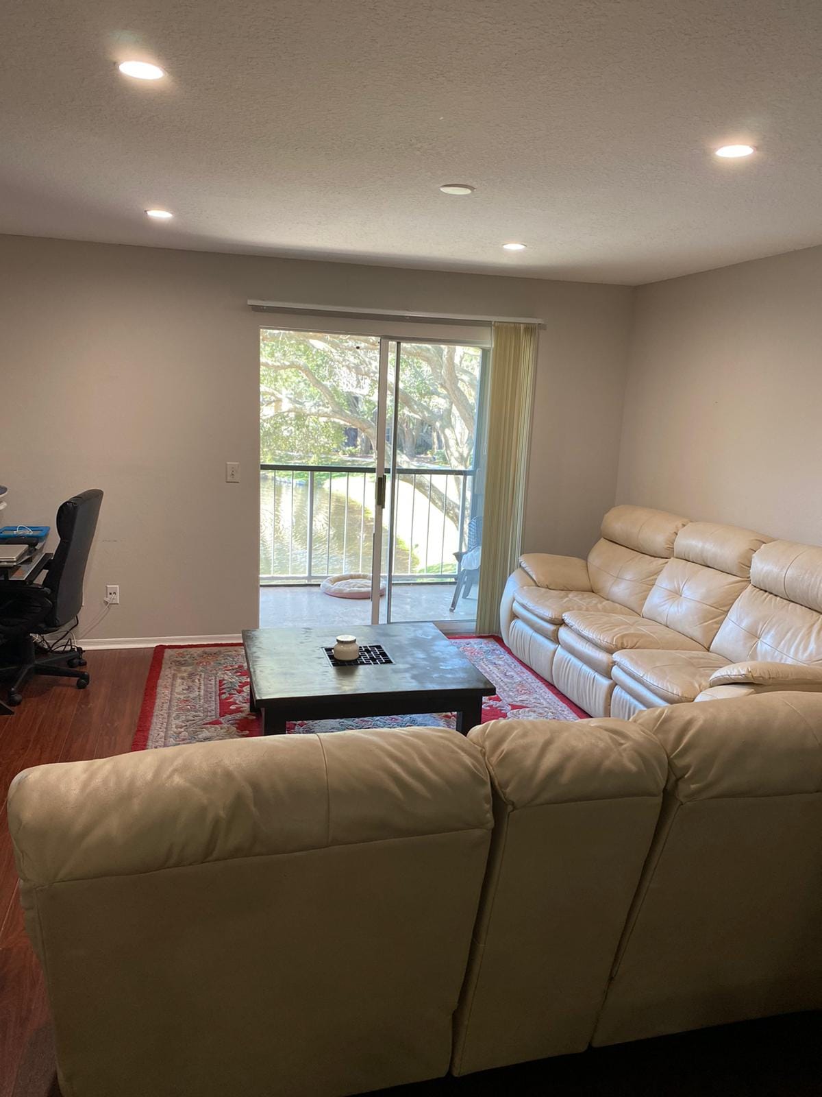 A light-filled living area features a leather sectional sofa arranged around a coffee table. A large window provides views of a tree-lined exterior, while a workspace is present in the corner, creating a functional and inviting environment.