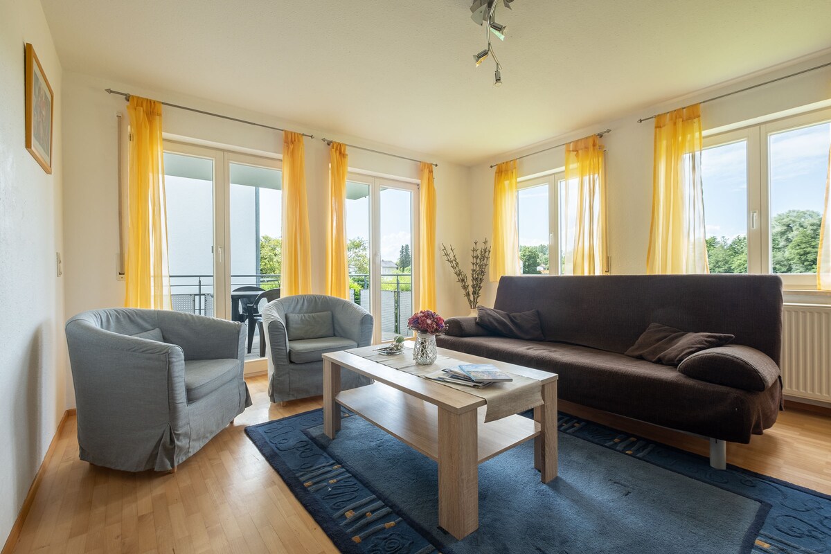 A spacious living area is enhanced with large windows that allow natural light to fill the space. Comfortable seating includes a dark sofa and two light blue armchairs. A wooden coffee table is centered on a blue area rug, further complemented by vibrant yellow curtains.