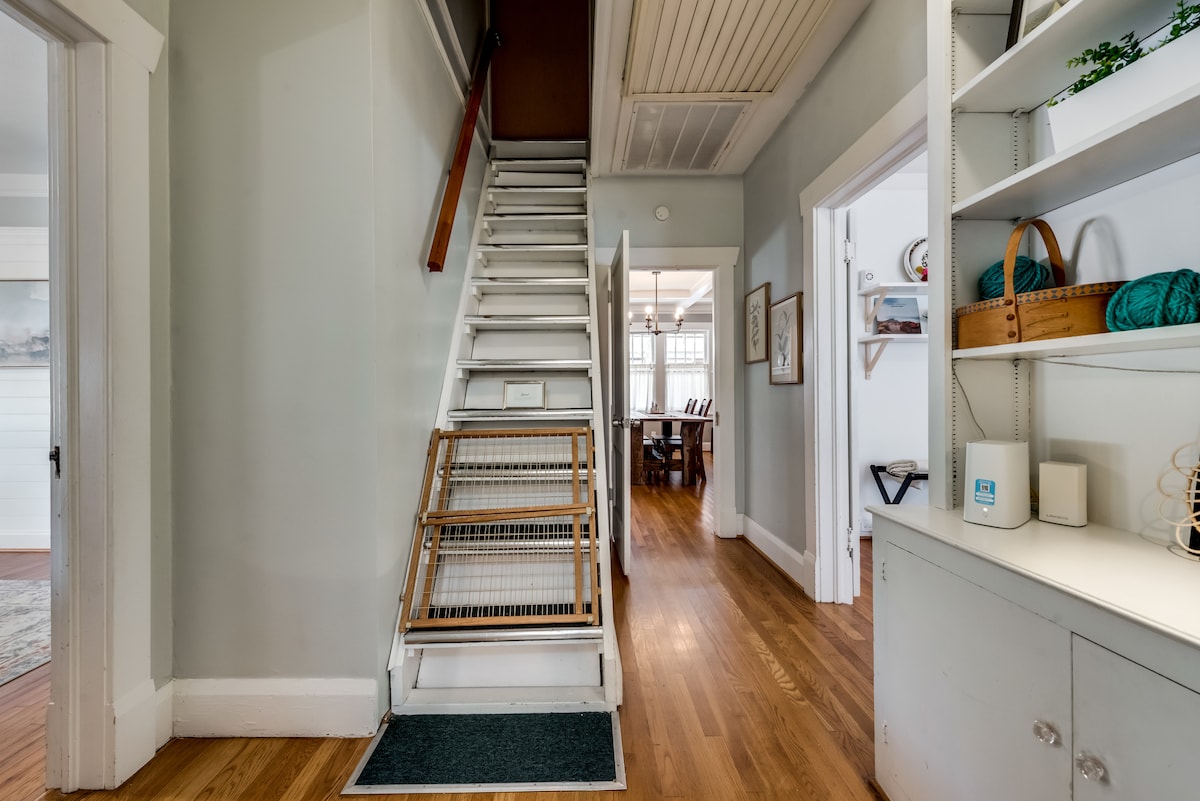 A well-lit hallway is depicted, featuring a wooden staircase leading to the upper level. Light-colored walls and polished hardwood floors enhance the spacious feel. Shelves with various items are positioned along one side, providing storage and decor. The end of the hallway opens to a dining area visible in the distance.