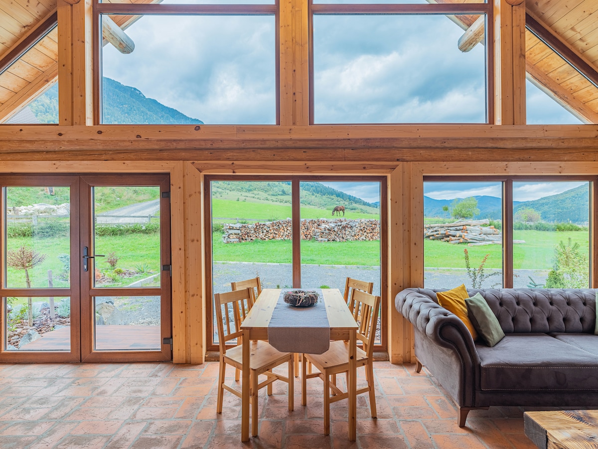 A spacious living area features large windows that frame a scenic view of green fields and distant mountains. A wooden dining table with four chairs is placed near the windows, while a comfortable sofa is positioned to invite relaxation.