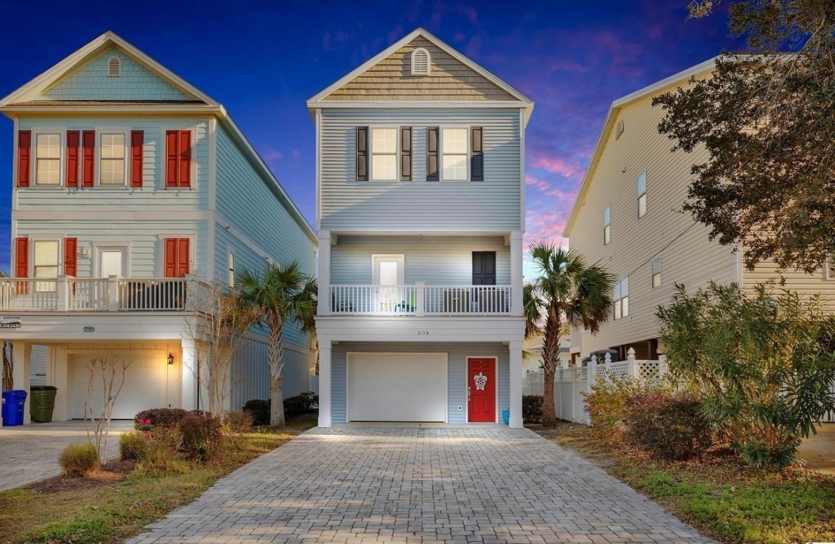 A two-story home is showcased with a welcoming red front door and a spacious driveway paved with stones. Tall palm trees are positioned alongside, with neighboring houses visible on either side. The home features balconies with white railings, framed by soft exterior lighting at twilight.