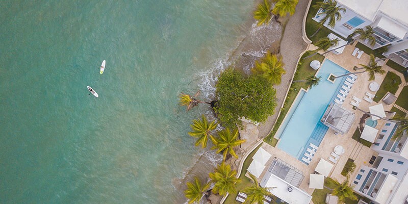 Aerial view of the resort showcases the shimmering turquoise water alongside the sandy shore. A pool surrounded by sun loungers and palm trees is visible, along with two paddleboarders in the water, highlighting the beachfront setting.