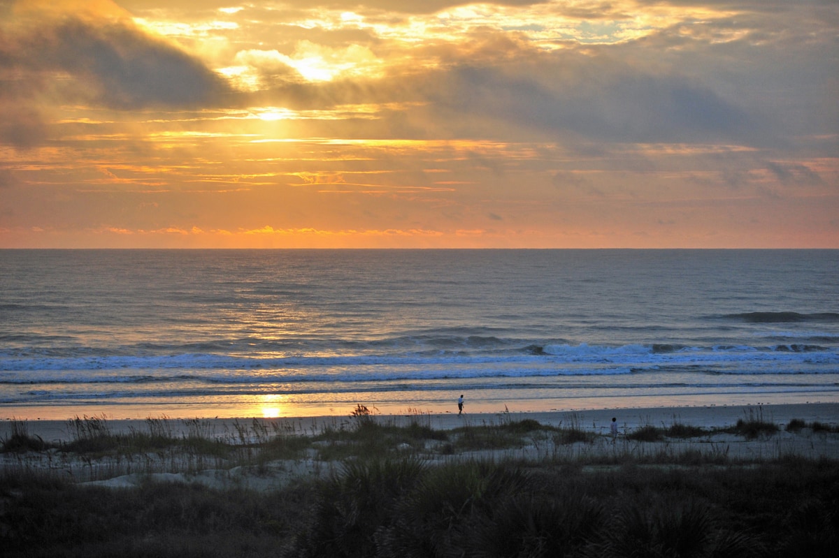 A serene beach landscape is depicted at sunrise, with gentle waves rolling onto the shore. A solitary figure can be seen walking along the water's edge, as the sky is filled with soft orange and golden hues. The scene captures the peaceful ambiance of the beachfront.