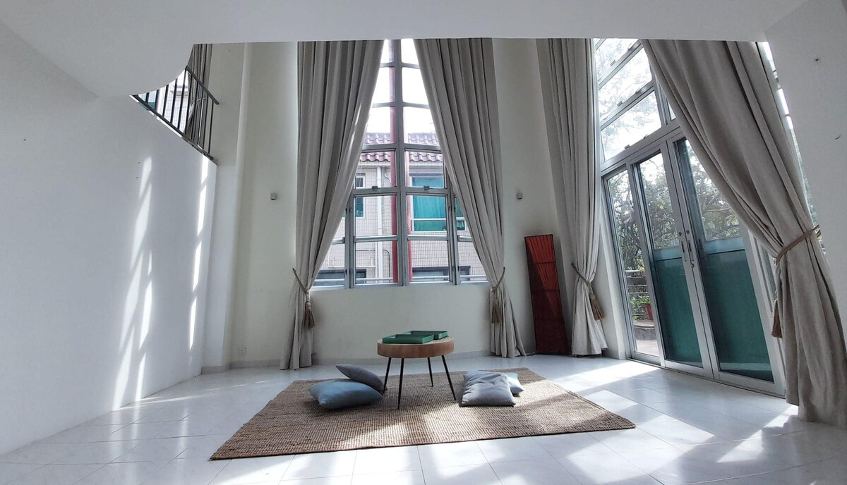 Tall windows invite ample natural light into a spacious living area, complemented by soft drapes. A circular wooden coffee table centers the space, surrounded by floor cushions on a woven rug. Clean white walls enhance the airy atmosphere, while greenery can be glimpsed through the glass doors.