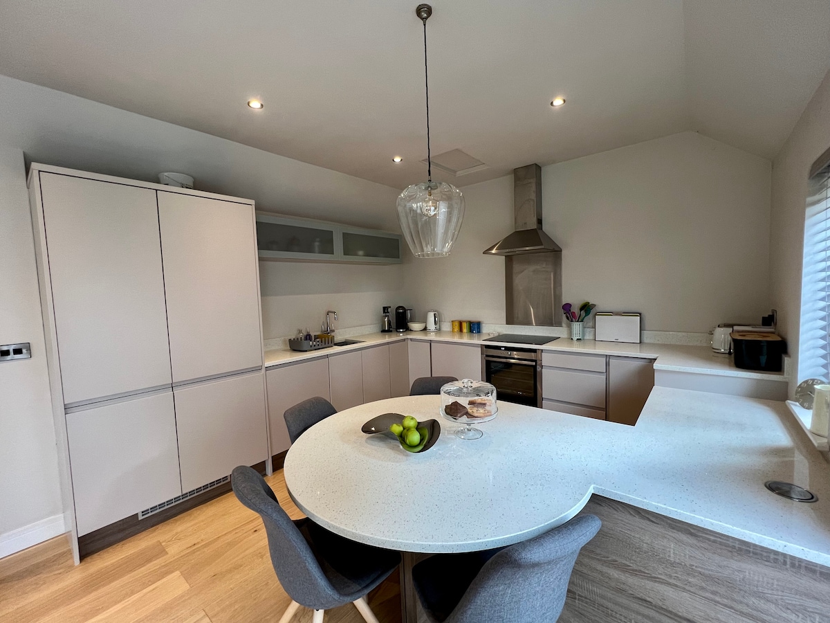 The kitchen features a modern design with sleek cabinetry and a spacious countertop. A round dining table with four chairs is positioned in the center, complemented by a large pendant light above. Kitchen appliances and utensils are neatly arranged, creating a functional space.