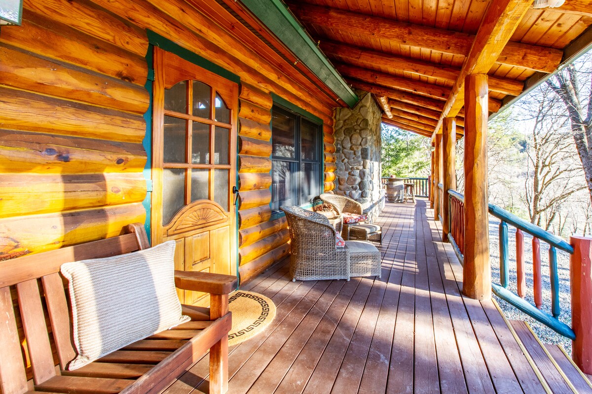 A spacious porch features a wooden floor with a combination of comfortable seating options, including wicker chairs and a wooden bench. Natural light filters in from large windows, highlighting the log cabin's rustic charm and surrounding wooded area.