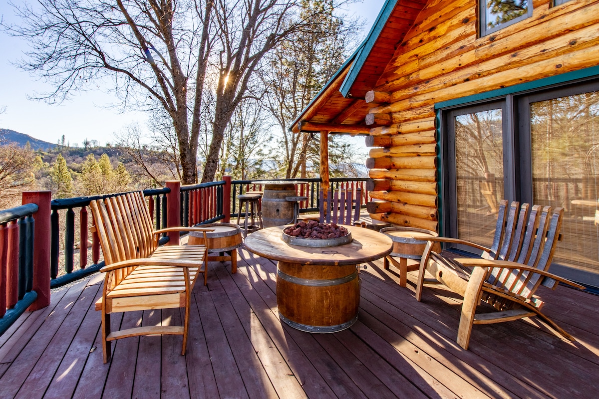 An outdoor deck features a round wooden table with a circular fire pit at its center. Surrounding the table are four rustic wooden chairs. The deck showcases a peaceful setting with trees and mountains visible in the background, illuminated by warm sunlight.
