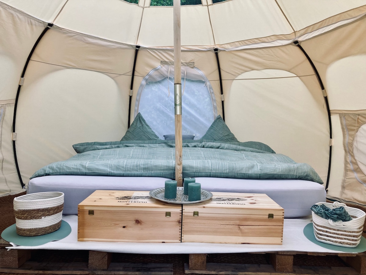 An inviting interior of a glamping tent features a spacious bed adorned with soft green linens. Natural light filters through a large window, while decorative baskets and wooden storage boxes create an organized and cozy atmosphere.