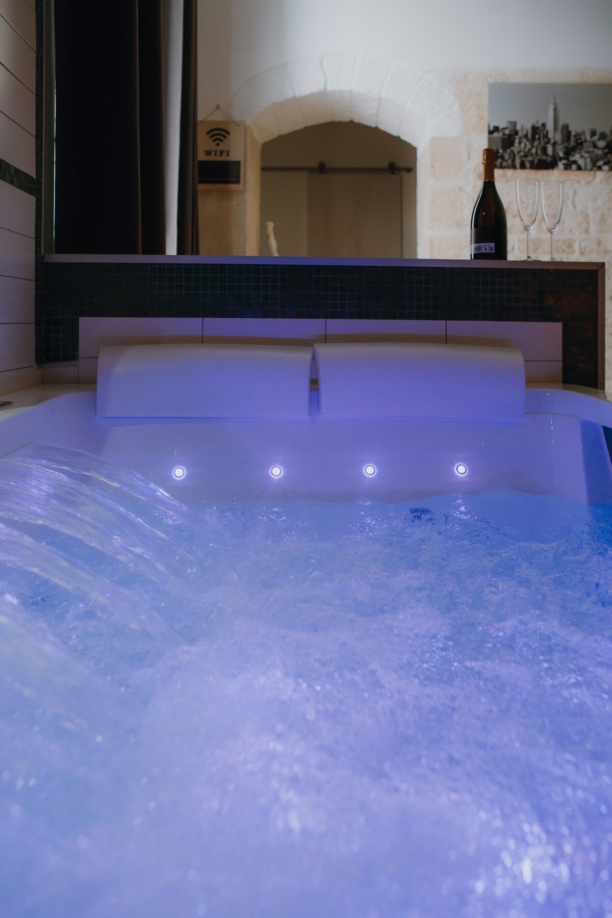 A spacious hydro-massage tub features gentle bubbles and soothing blue lighting. The tiled walls and sleek design create a modern aesthetic. A bottle and glasses are visible on a ledge nearby, enhancing the relaxation space in the inviting ambiance of the room.