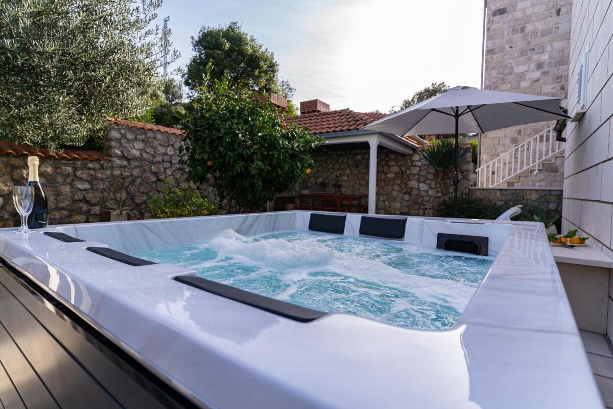 A spacious outdoor jacuzzi is nestled in a private garden, surrounded by lush greenery and built stone walls. An umbrella provides shade over one side, while a table can be seen nearby. Bubbles gently rise in the water, ready for relaxation.