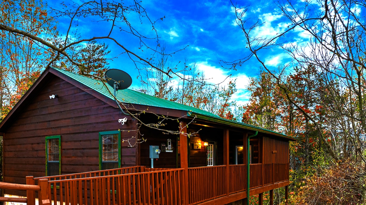 The log cabin features a metal roof, surrounded by trees showcasing seasonal colors. A spacious deck with wooden railings extends from the front, providing an inviting entrance. Large windows offer glimpses of the interior, while the sky adds a vibrant backdrop.