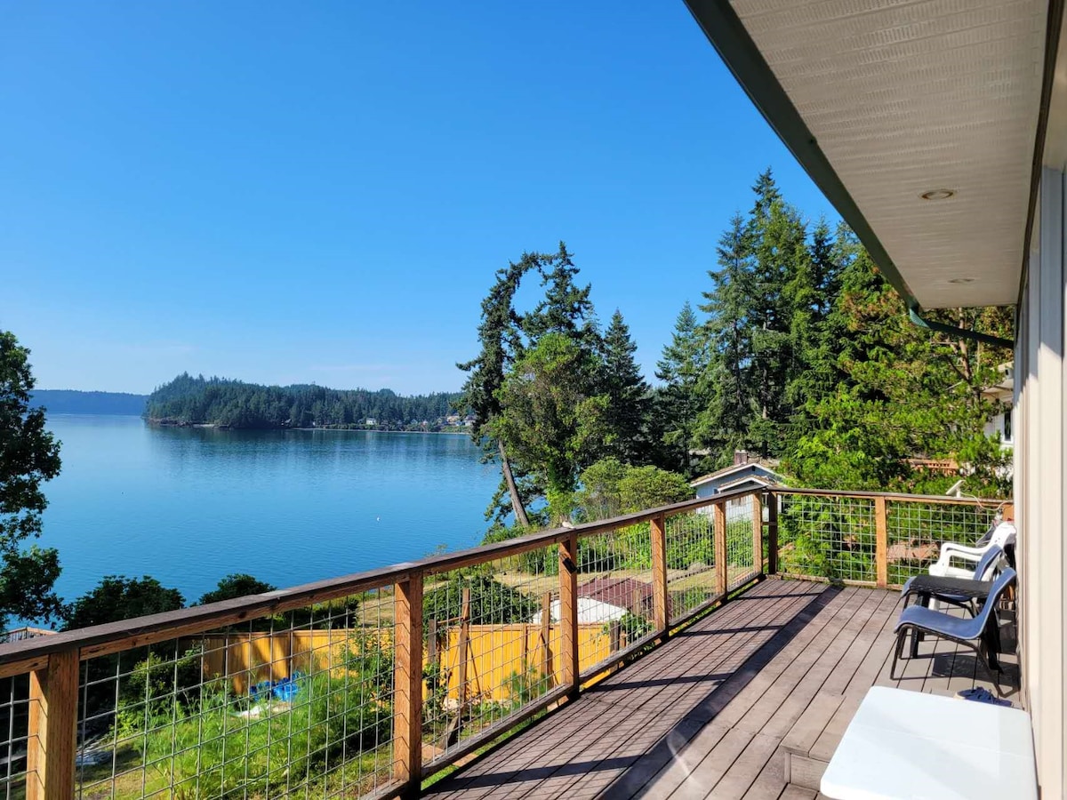 A spacious deck overlooks a calm body of water, framed by lush greenery and distant hills. Comfortable lounge chairs are positioned for enjoying views of the serene landscape under a clear blue sky.