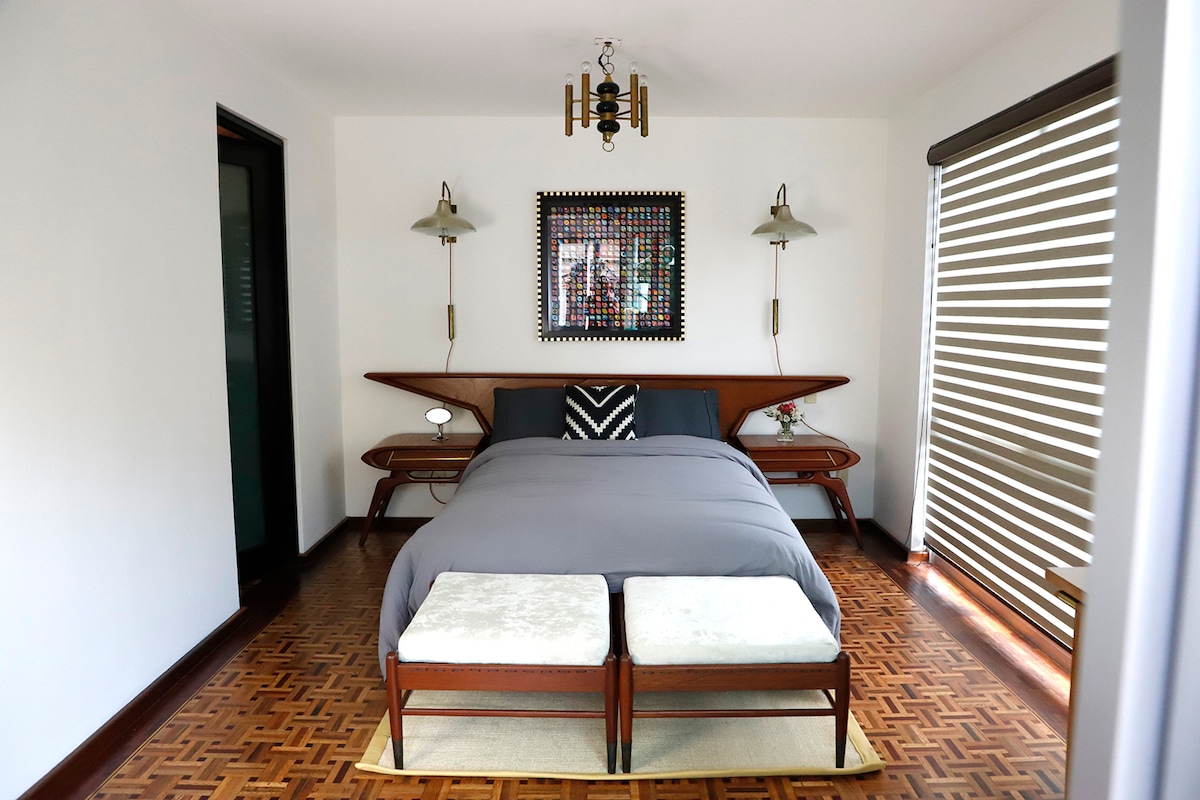 A well-appointed bedroom is presented, featuring a spacious bed dressed in gray bedding. Two upholstered benches sit at the foot of the bed. Wall-mounted lamps provide illumination, and an artwork is displayed above the bed. Large windows with blinds bring in natural light.