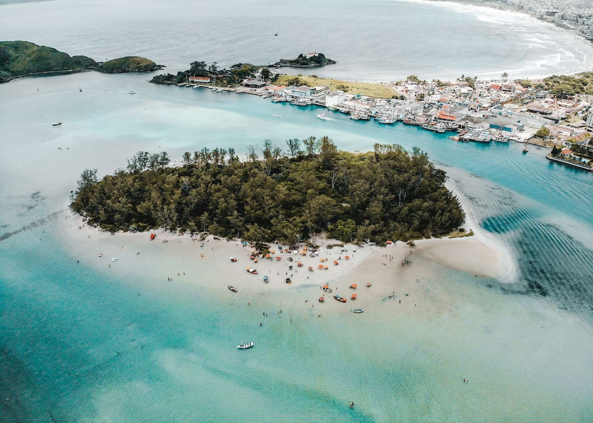 An aerial view of a small island surrounded by calm turquoise waters is presented. The sandy beach is dotted with sunbathers and colorful umbrellas, while small boats are anchored nearby. In the background, the coastline of a bustling area can be seen.