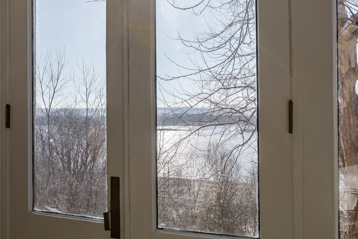 Two glass-paneled doors frame a view of the Mississippi River, with sunlight reflecting off the water's surface. Trees with bare branches are visible in the foreground, indicating a seasonal transition.
