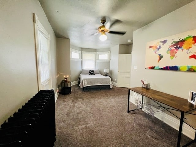 A well-lit room features a spacious bed positioned against the wall, accompanied by a decorative world map hanging above a wooden table. Natural light filters through bay windows, and a ceiling fan provides airflow. A soft carpet covers the floor, enhancing comfort.