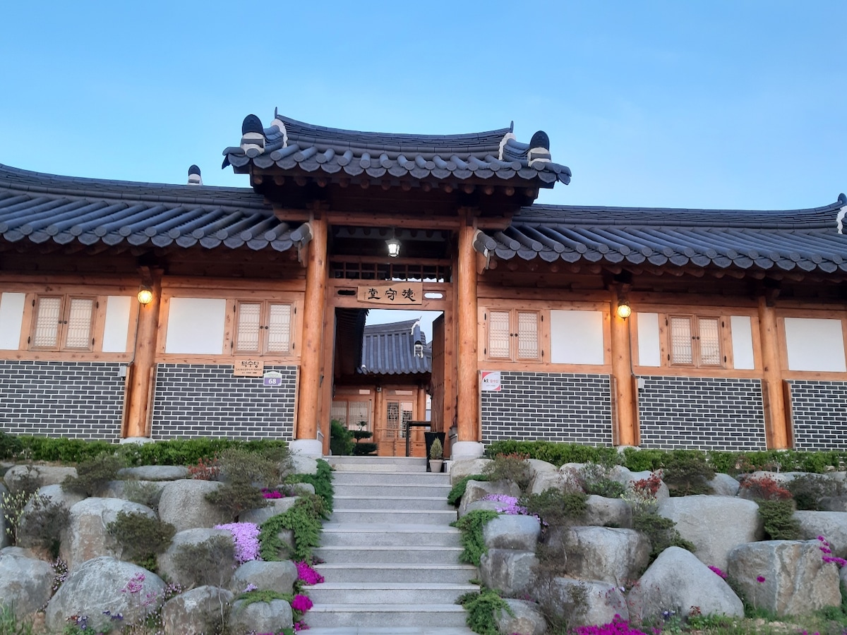 A traditional Korean house showcases elegant architecture, highlighted by its wooden beams and tiled roof. Lush greenery and colorful flowers surround the entrance, creating a serene environment. Soft lighting enhances the inviting atmosphere of the exterior during twilight.