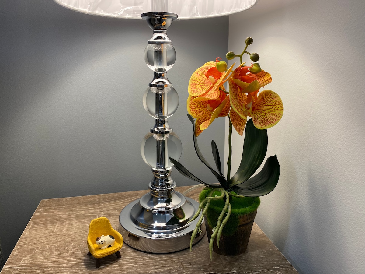 A stylish lamp with a clear base and white shade is positioned beside a small, vibrant yellow chair. An artificial orchid with bright orange and yellow petals adds a pop of color, resting in a green pot on the wooden side table.
