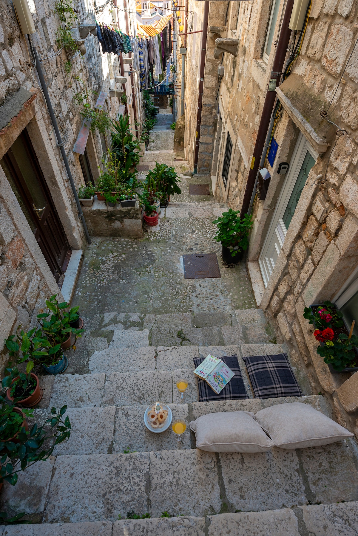 A narrow stone alleyway is lined with traditional buildings, featuring potted plants and hanging laundry. Steps lead down to a small seating area with pillows and a blanket, accompanied by a book and two drinks on a tray. Sunlight filters through, illuminating the vibrant scene.
