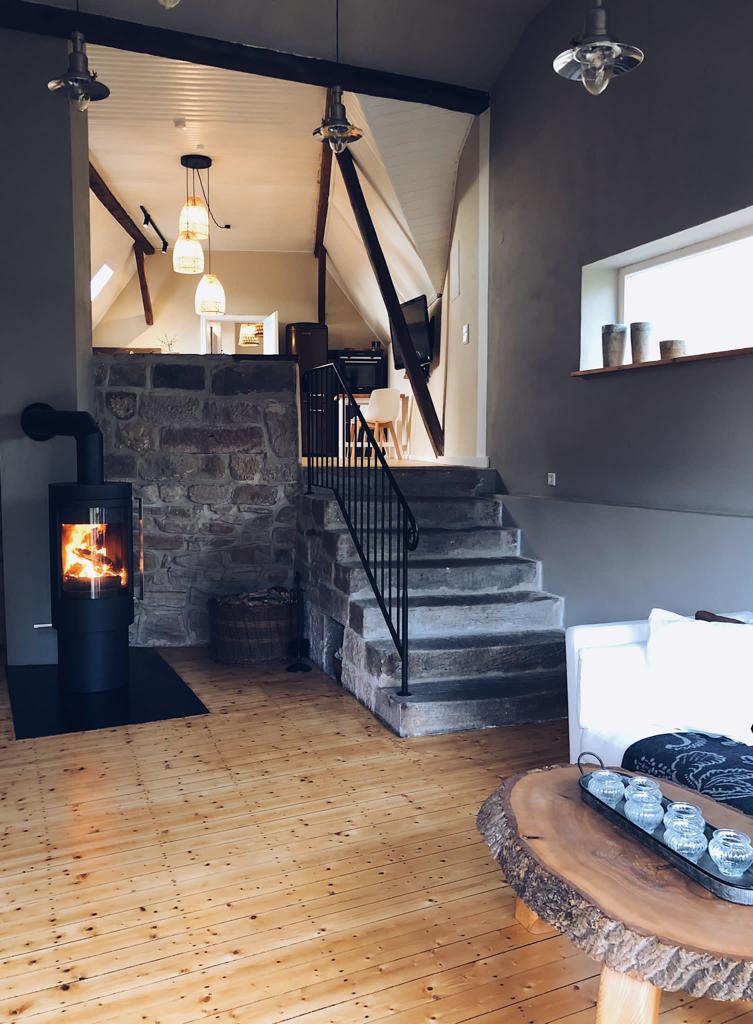 A spacious loft area is featured with wooden flooring and exposed beams. A sleek fireplace stands near the stone wall, providing warmth. A staircase leads to the upper level, while modern lighting fixtures hang from the ceiling, softly illuminating the space.