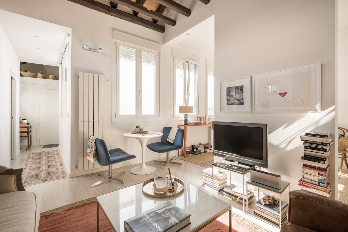 A bright living area features a coffee table at the center, surrounded by two modern chairs and a sofa. Light enters through multiple windows, illuminating stylish decor, including framed artwork on the walls and a well-organized bookshelf. A soft rug adds warmth to the space.