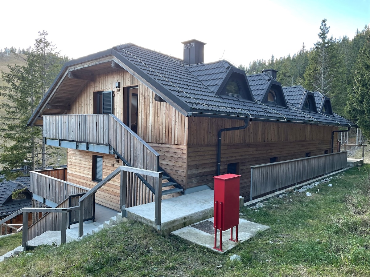 A wooden structure is nestled among trees, showcasing a sloped roof and multiple dormer windows. An exterior staircase leads to the upper level, while a red mailbox stands beside a concrete path, enhancing the natural surroundings.