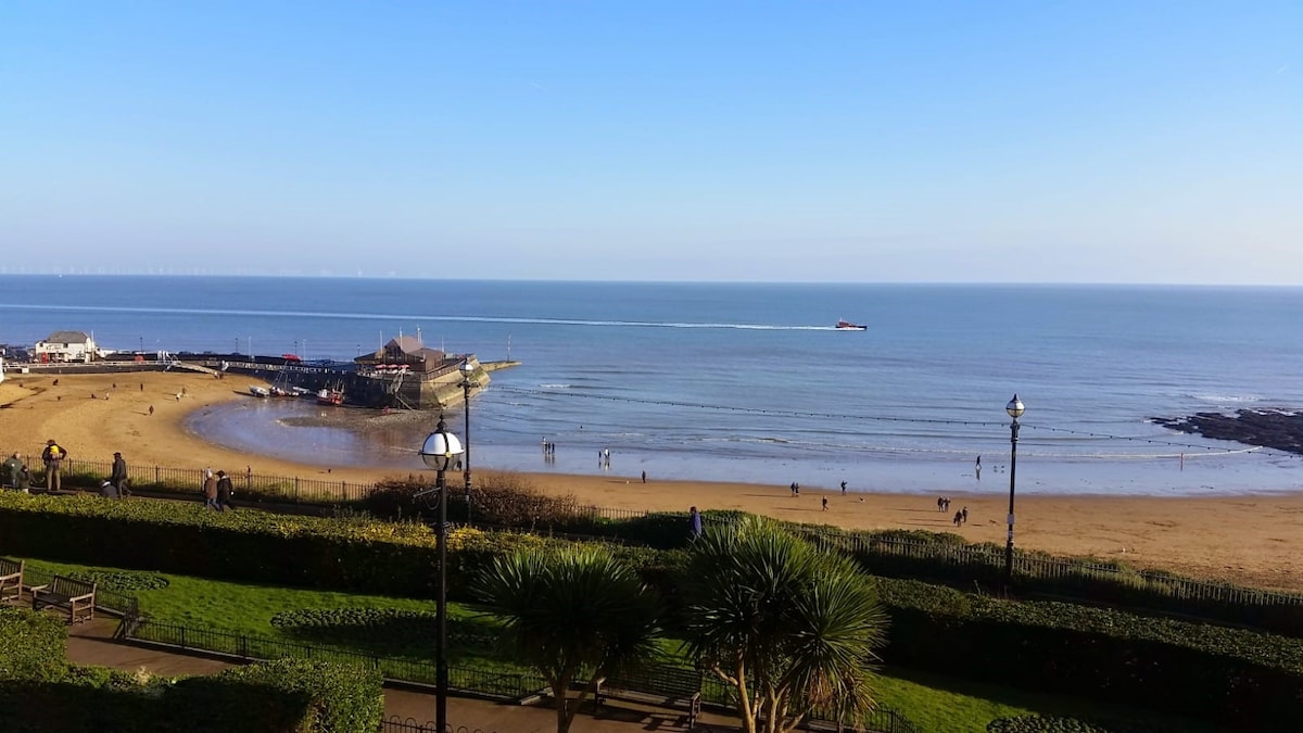 A panoramic view of Viking Bay is captured, revealing a sandy beach with gentle waves lapping at the shore. People can be seen strolling along the beach and pier. Lush greenery and palm-like plants are present in the foreground, framing the scenic seascape.