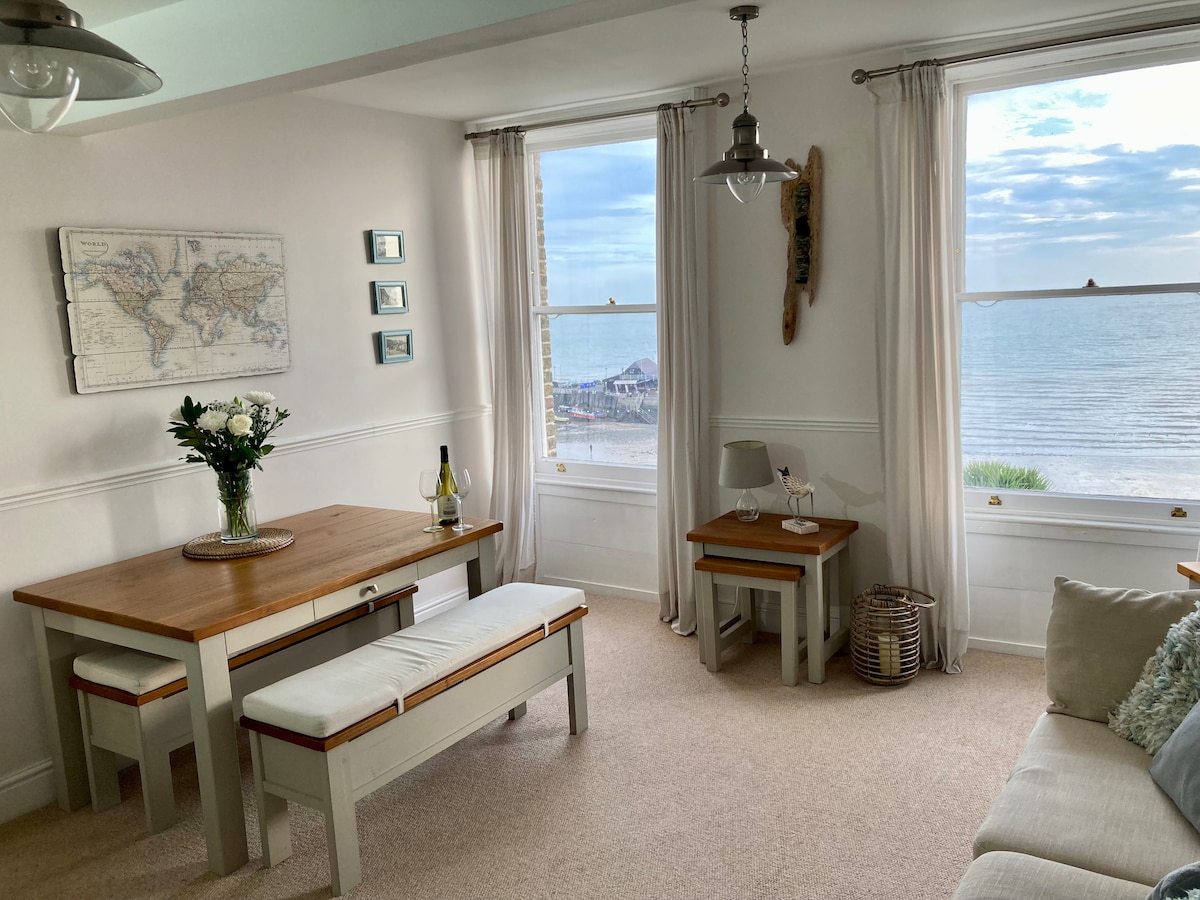 The dining area features a wooden table with two bench seats and a side table, accompanied by a vase of flowers. Large windows provide a view of the sea and the nearby beach, with soft natural light enhancing the space.