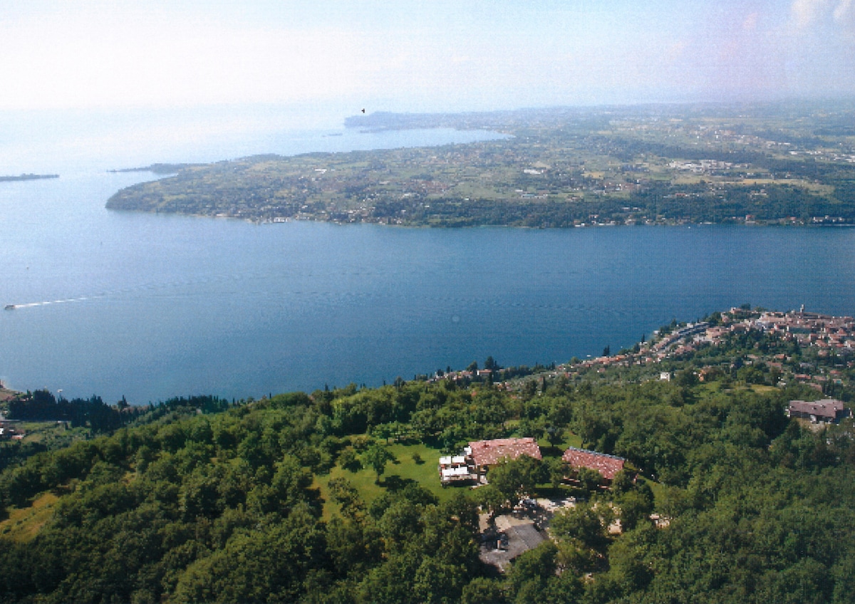 An expansive view of the Garda Lake is captured, showcasing its tranquil blue waters surrounded by lush greenery and rolling hills. The landscape features small villages along the lakeshore, with the farmhouse prominently situated on a terraced property, blending harmoniously with its natural surroundings.