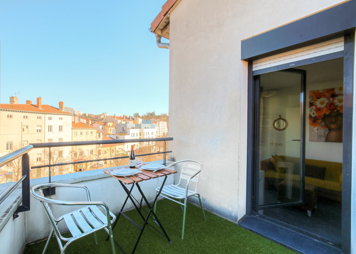 A small balcony is visible, featuring a table set for two and two chairs. An unobstructed view of the surrounding buildings and green hills is offered. The door leading to the interior is partially open, inviting natural light into the space.