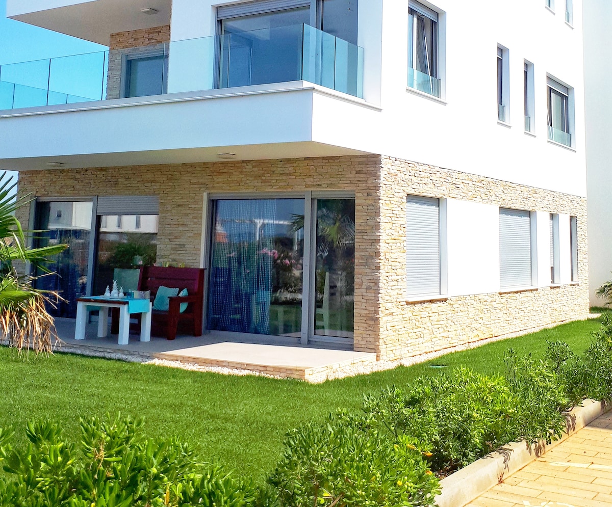 The exterior of the apartment showcases a modern design with a stone facade and large glass doors opening to a patio. A small table and chairs are positioned on the terrace, surrounded by a well-maintained grassy area and garden plants.