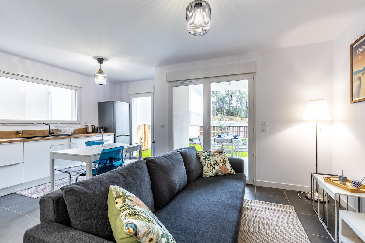 A modern living area features a comfortable dark sofa adorned with decorative pillows. Natural light streams through the large sliding doors, leading to an outdoor space. A simple dining table with blue chairs and a sleek kitchen are visible in the background.