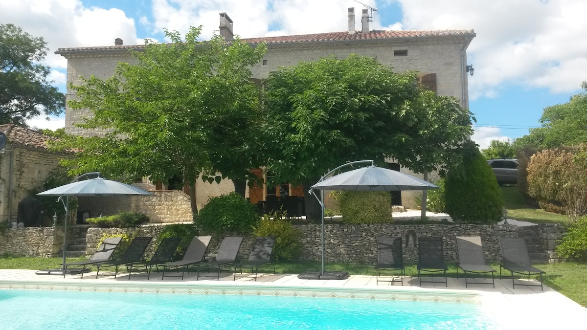 A serene outdoor scene features a spacious swimming pool surrounded by lush greenery and eight lounge chairs. Two umbrellas provide shade beside the pool, while a charming stone building with large windows and a sloping roof is visible in the background. Fluffy clouds drift across the blue sky.