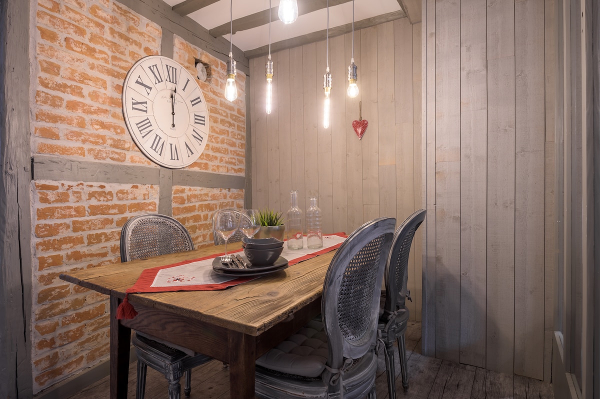 A dining area features a rustic wooden table accompanied by four grey, woven chairs. A large wall clock is mounted above, adding character to the space, while pendant lights hang from the ceiling, providing illumination. Exposed brick and wooden paneling enhance the cozy ambiance.