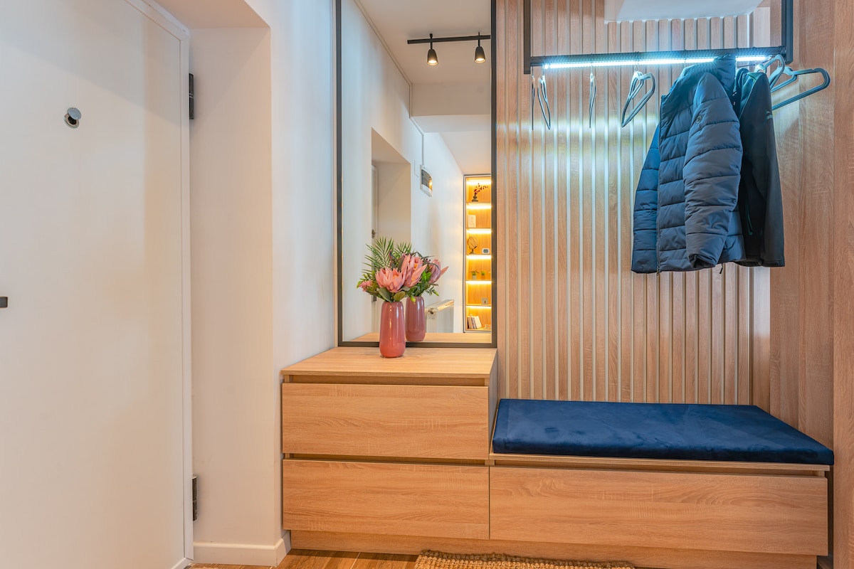 A welcoming entryway features a wooden bench and storage drawers. A coat rack displays a dark jacket, while a vase with fresh flowers adds a touch of color. Soft lighting from overhead fixtures enhances the cozy atmosphere.
