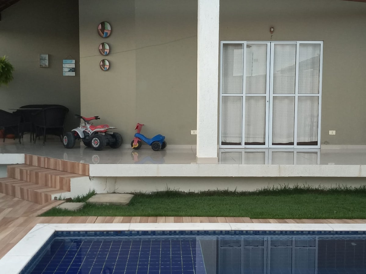 An outdoor area features a private pool, complemented by a patio and steps leading to a grassy edge. Two children's toy vehicles are displayed beside the pool, and a covered seating area is visible in the background, framed by large glass doors.