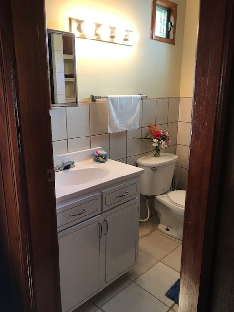 The bathroom features a light-colored tile design with a spacious vanity and a large mirror above it. A small vase of flowers adds a touch of color, while the toilet is positioned beside the vanity. Soft lighting is provided by a fixture above the mirror.
