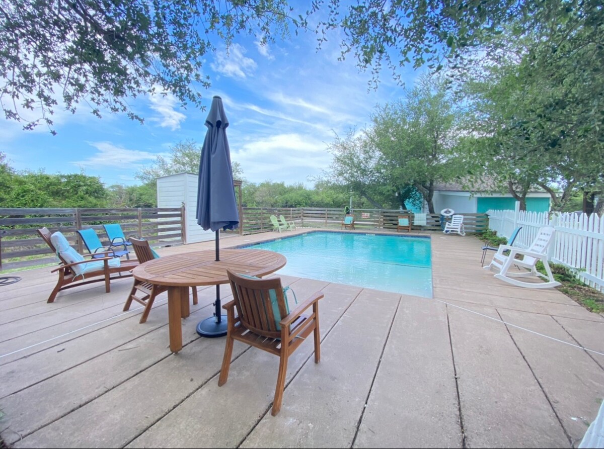 A community pool is surrounded by a spacious deck featuring several seating areas, including lounge chairs and a dining table with an umbrella. Lush trees provide shade while a white fence encircles the pool area, enhancing privacy and tranquility.