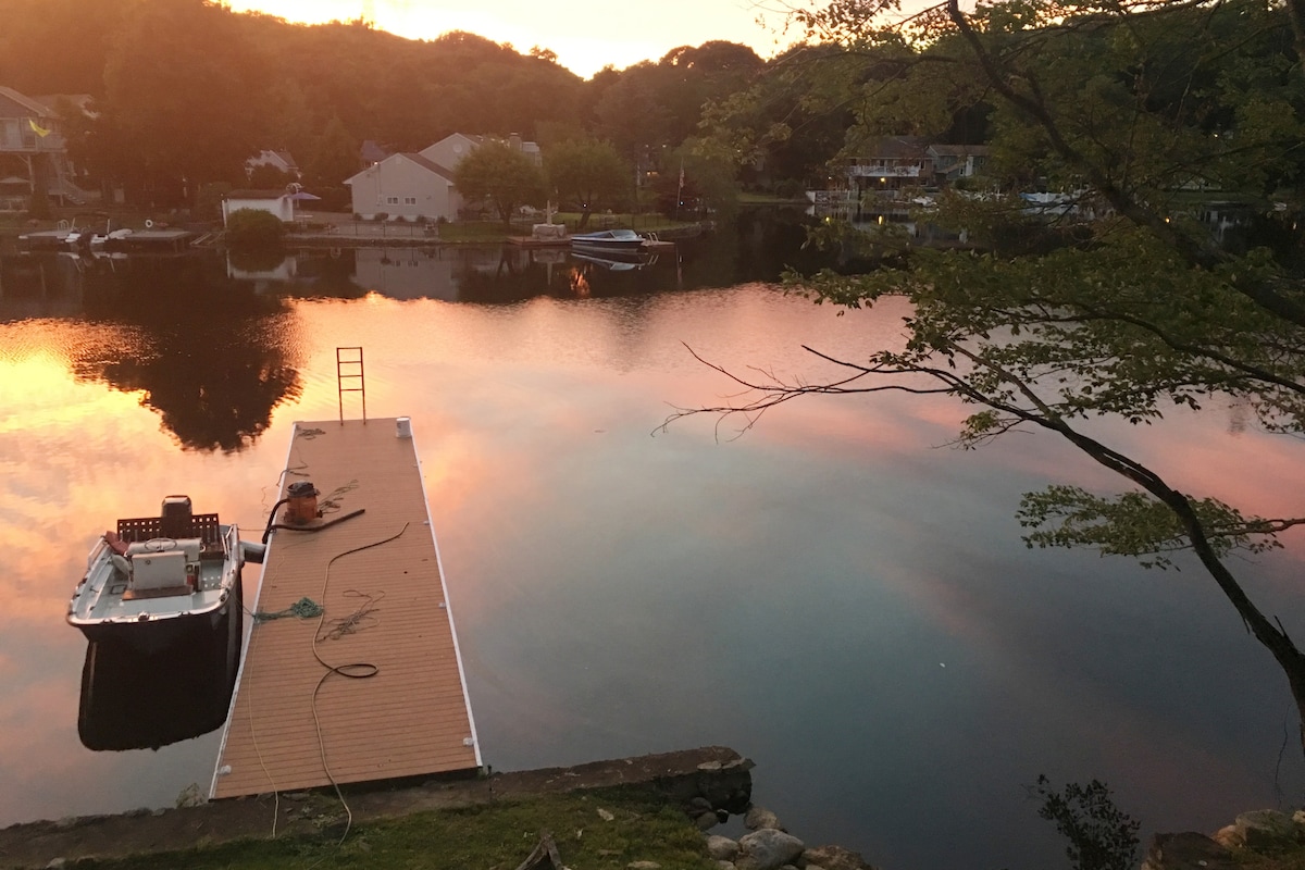 A tranquil lakefront scene is captured at sunset, with vibrant colors reflecting on the water. A stationary dock extends into the lake, accompanied by a small rowboat tied to its side. Lush greenery surrounds the area, enhancing the peaceful ambiance.