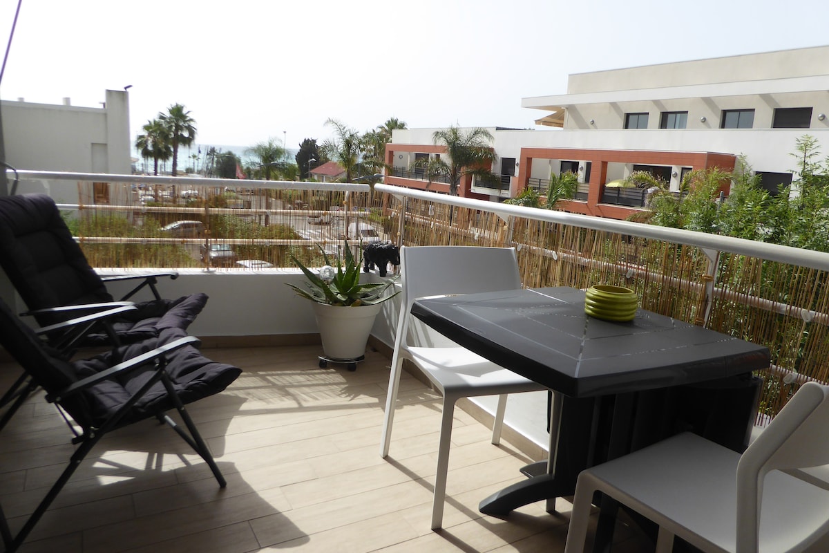 A terrace with a table and chairs overlooks a scenic view that includes greenery and glimpses of the sea. A black lounge chair is positioned nearby, and potted plants add a touch of nature to the space.