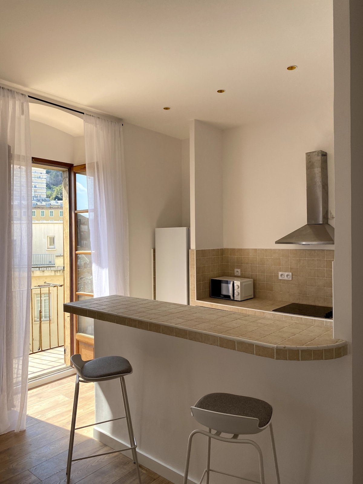 A light-filled kitchen area is seen featuring a bar with two stools and a minimalist design. A microwave is positioned on the counter, and a window allows for natural light to flood the space, leading to a charming balcony with open views.