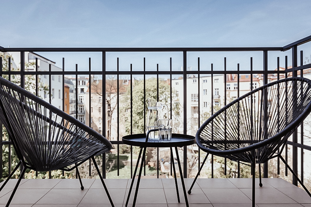 A private balcony is featured with two modern chairs and a small table, showcasing a view of the surrounding buildings and trees. Natural light fills the space, creating a bright atmosphere, while the railing provides safety and enhances the open feeling.