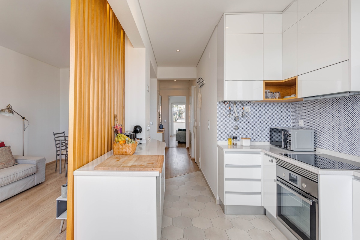 A modern kitchen area features sleek white cabinetry and a patterned backsplash. A wooden countertop is positioned near a fruit bowl, enhancing the functional space. The open layout connects to a nearby living area, with light streaming in through large windows, contributing to a bright atmosphere.
