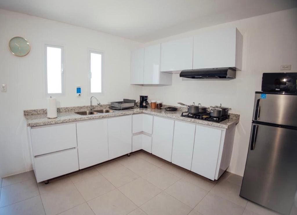 A modern kitchen features sleek white cabinetry and a spacious countertop. A stainless steel refrigerator stands next to a stove, with pots and utensils arranged neatly on the surface. Light streams in through two small windows, illuminating the functional space.
