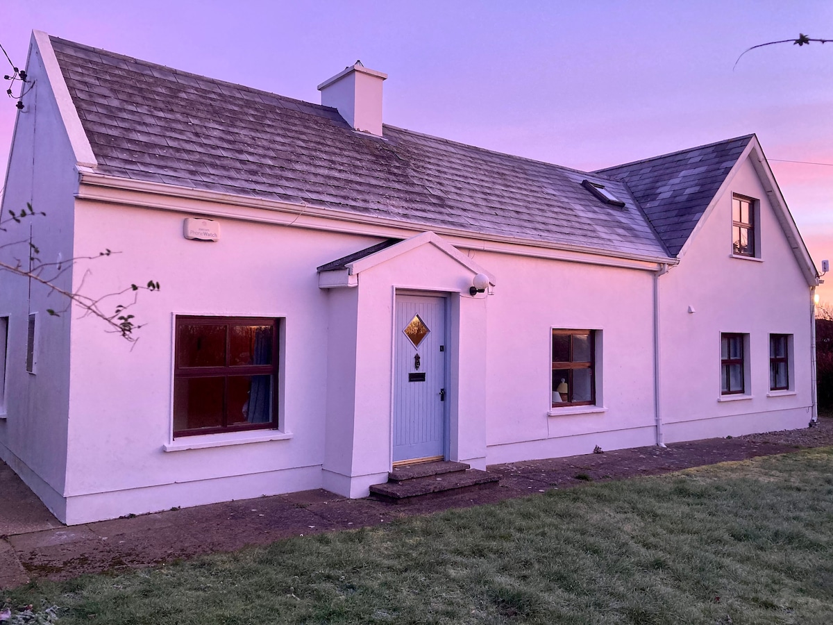 A white cottage with a gabled roof is set against a soft purple sky. The front features a centrally located blue door, flanked by large windows. The ground is grassy, with a clear view of the structure's clean lines and simple design.