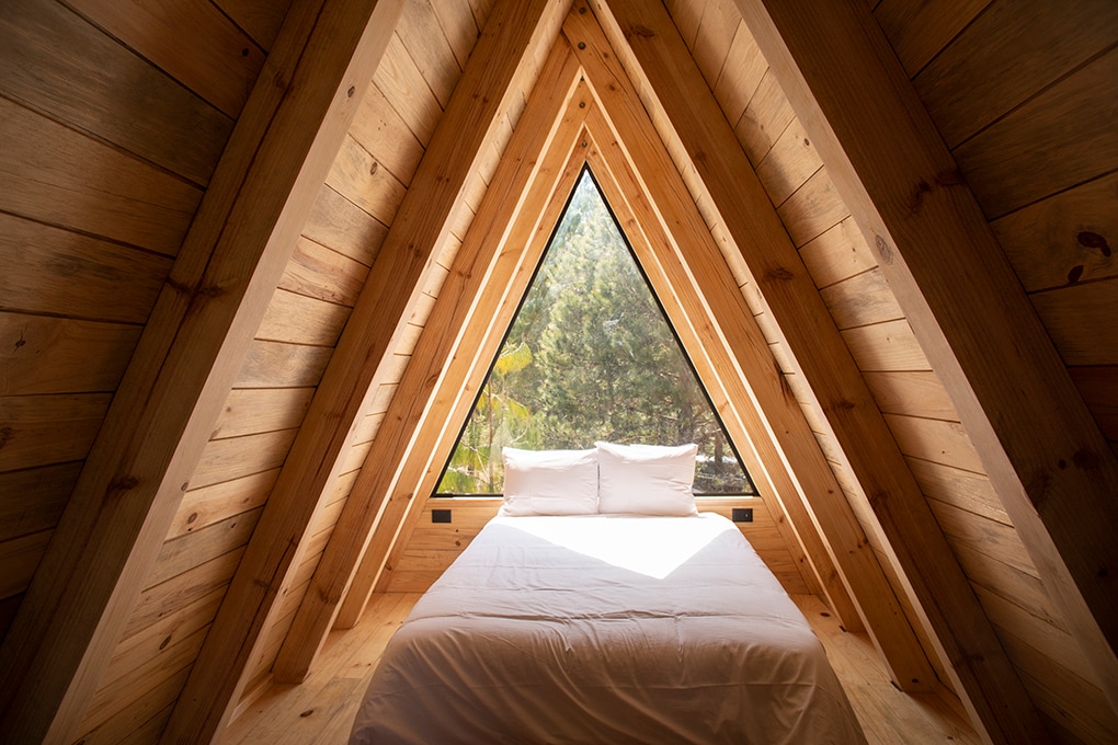 The interior of a wooden chalet features a spacious bed positioned beneath a striking triangular window. Natural light pours in, highlighting the wooden beams and walls, creating a cozy ambiance. Outside the window, greenery suggests a serene forest setting.
