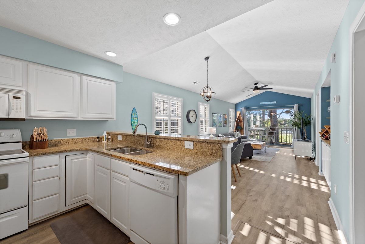 A modern kitchen is shown with white cabinetry and granite countertops, featuring essential appliances including a microwave and dishwasher. Natural light enters through multiple windows, illuminating the open layout that connects to the living area, which includes seating and a view of the outdoor space.