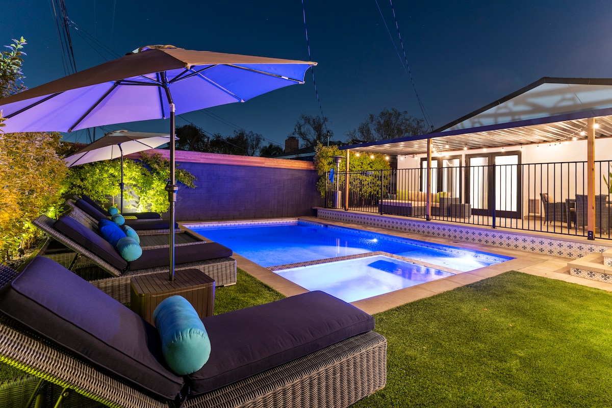 A serene outdoor space features a sparkling pool illuminated by ambient lighting. Comfortable lounge chairs are arranged beside the pool, each with a blue cushion. A covered seating area extends to the side, surrounded by lush greenery, creating an inviting environment for relaxation.