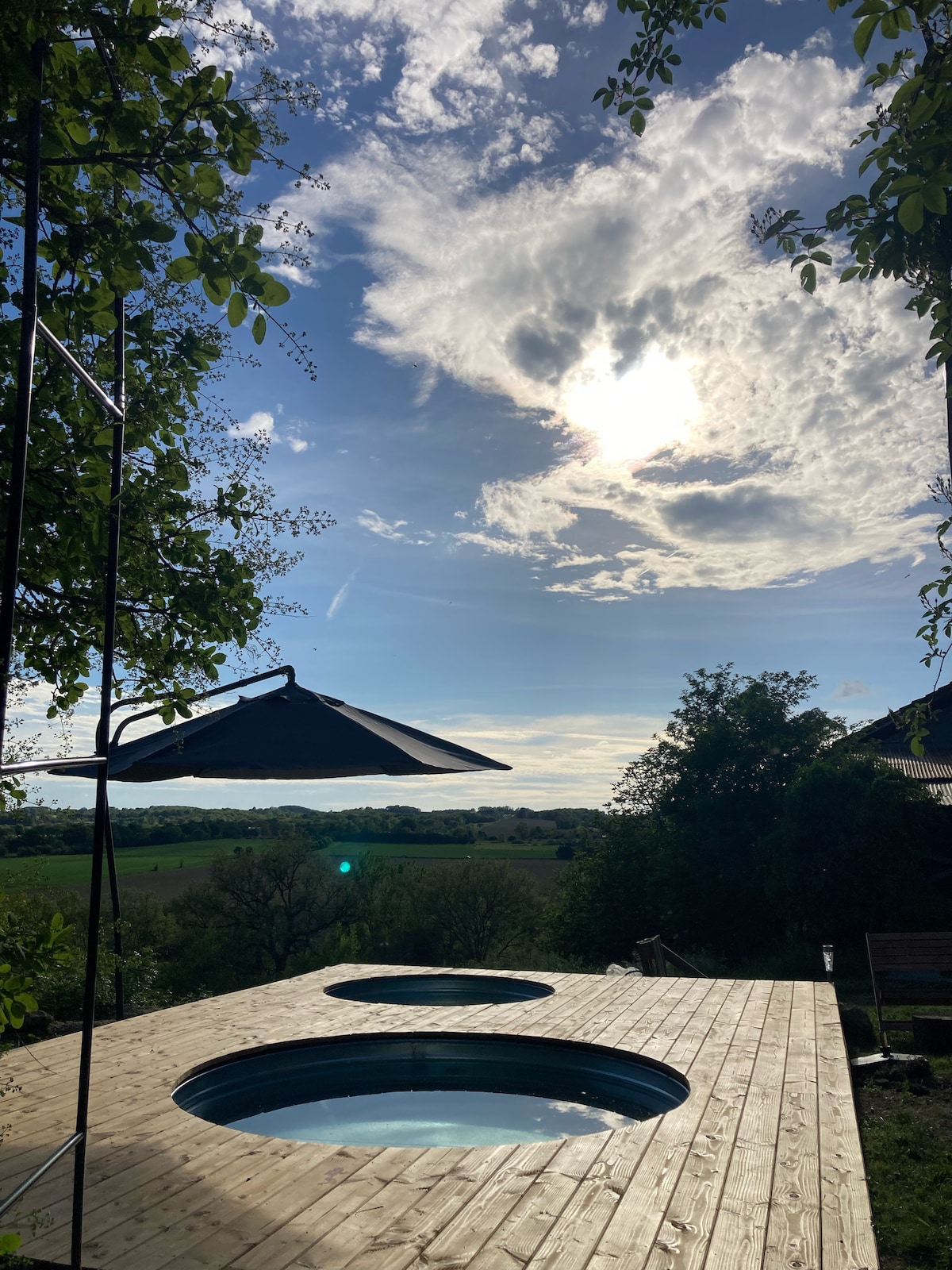 Two round soaking tubs are set on a wooden deck surrounded by greenery. A shade umbrella stands nearby, and the sky is adorned with clouds and sunlight, casting a serene glow over the landscape.
